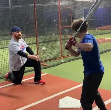 Coach kneeling to soft toss to a young hitter in an indoor training cage